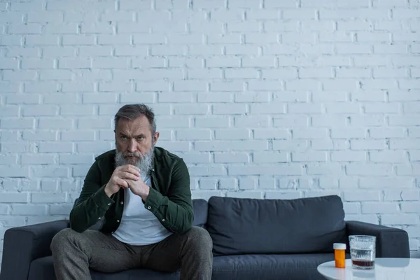 Deprimido hombre mayor con el pelo gris sentado en el sofá cerca de la botella con medicamentos y un vaso de whisky en la mesa de café - foto de stock