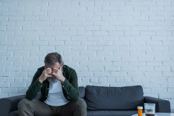 Deprimido hombre mayor con el pelo gris sentado en el sofá cerca de la botella con medicamentos y un vaso de whisky en la mesa de café - foto de stock