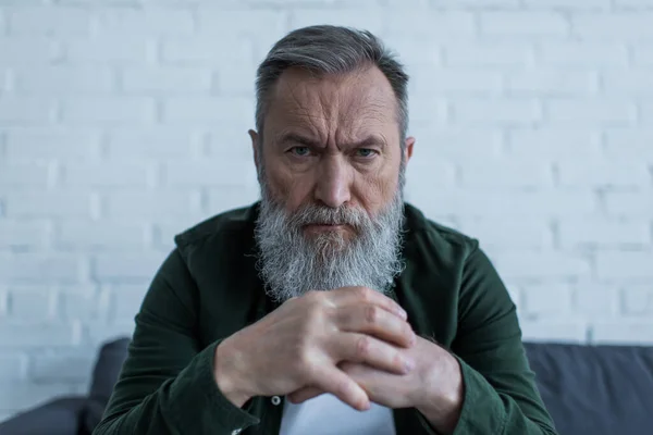 Serio hombre mayor con barba frunciendo el ceño mientras sufre una crisis y mirando a la cámara - foto de stock