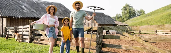 Famiglia con ciotola e rastrelli sorridente alla telecamera vicino al recinto in fattoria rurale, banner — Foto stock