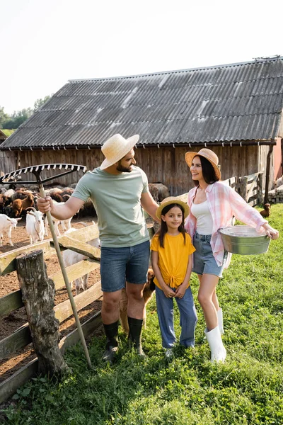 Granjeros felices en sombreros de paja hablando cerca de hija y corral en la granja - foto de stock