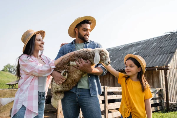 Happy farmer in straw hat holding lamb near daughter and wife on farm - foto de stock