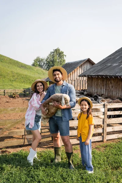 Agriculteur avec agneau dans les mains souriant à la caméra avec la famille à la ferme dans la campagne — Photo de stock