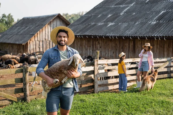 Granjero feliz con cordero mirando la cámara cerca de la familia y el perro de ganado en la granja - foto de stock