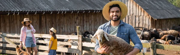 Agriculteur en chapeau de paille tenant agneau et souriant à la caméra près de la famille au corral, bannière — Photo de stock