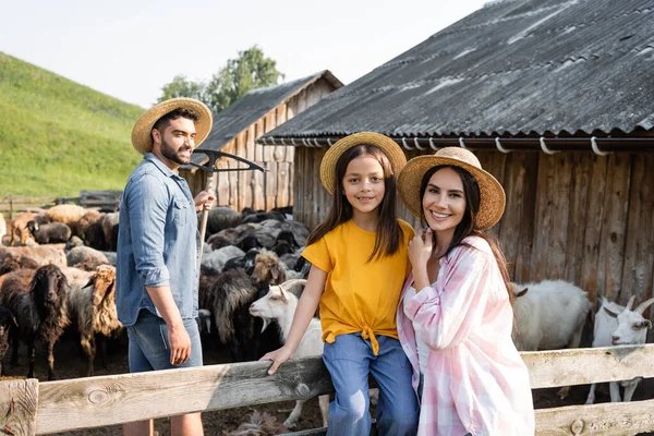 Mom and daughter in straw hats smiling at camera near farmer in corral - foto de stock