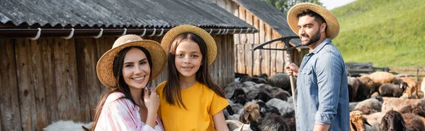 Happy family in straw hats looking at camera near sheep flock in corral, banner — Stock Photo
