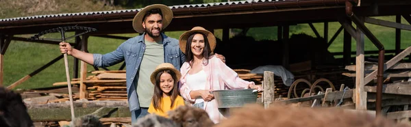 Família feliz de agricultores em chapéus de palha perto curral de gado em primeiro plano borrado, bandeira — Fotografia de Stock