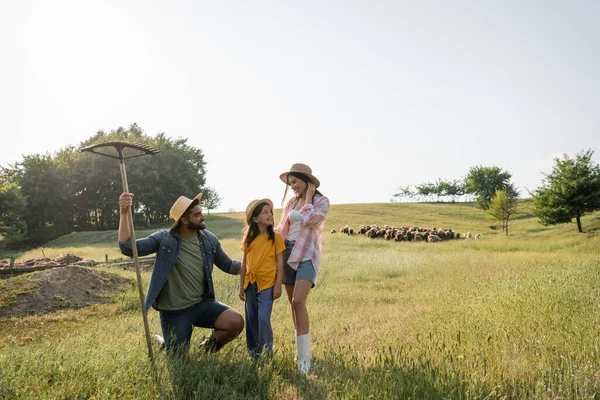 Farmer holding rakes near smiling family and herd grazing in pasture — Foto stock