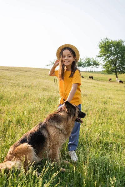Child in straw hat smiling at camera near dog and herd grazing in pasture — Photo de stock