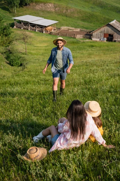 Granjero feliz en sombrero de paja caminando hacia la familia sentado en un prado pintoresco - foto de stock