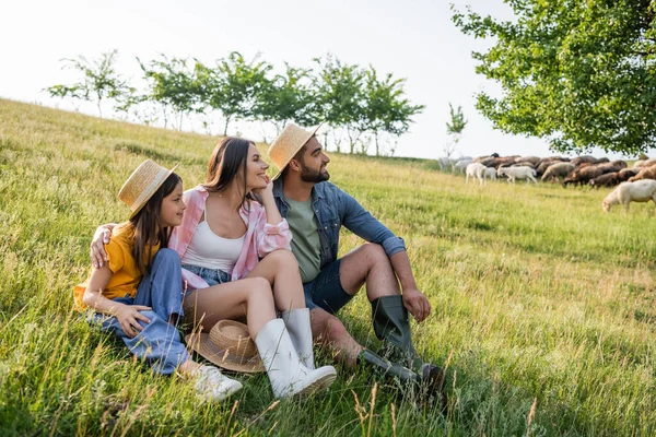 Famiglia gioiosa in cappelli di paglia seduto in pascolo erboso e guardando altrove — Foto stock