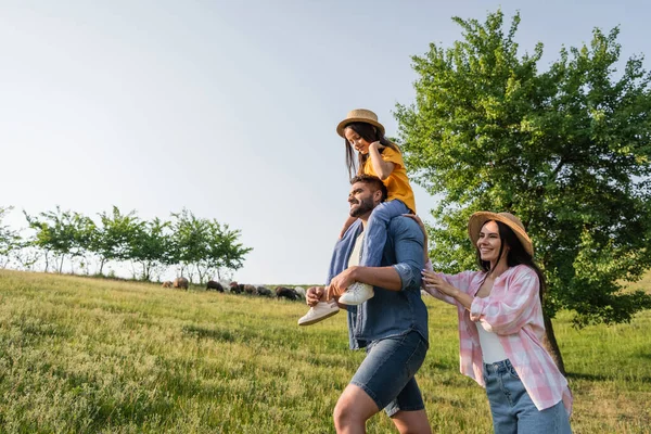 Smiling farmer piggybacking daughter while walking with wife in green meadow — Stock Photo