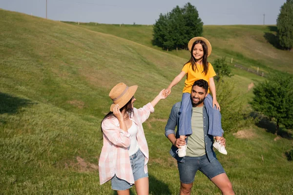 Happy man piggybacking daughter near wife while walking on green hills — Photo de stock