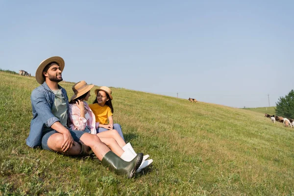 Glücklicher Bauer schaut weg, während er bei Familie auf dem Feld unter blauem Himmel sitzt — Stockfoto