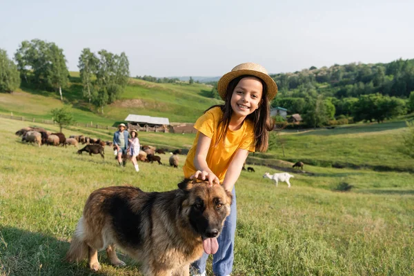 Menina alegre olhando para a câmera perto de cão de gado e pais rebanho pastoreio no fundo borrado — Fotografia de Stock