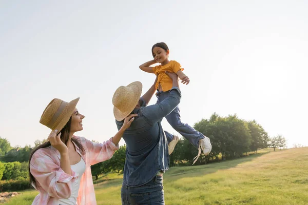 Man in straw hat raising up cheerful daughter near happy wife — Stock Photo