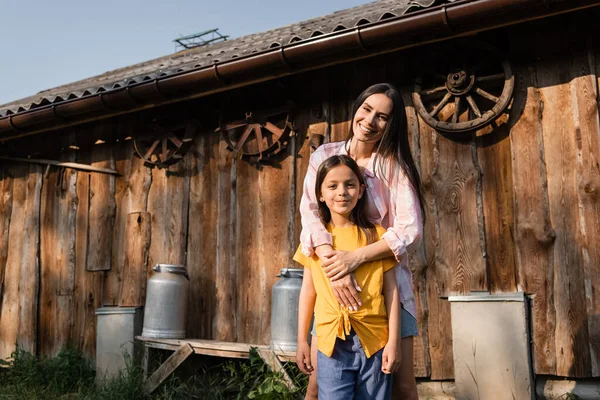Happy woman hugging daughter and smiling at camera near wooden barn — Photo de stock