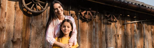 Cheerful woman embracing daughter near wooden barn on farm, banner — Stock Photo
