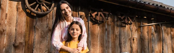 Cheerful woman embracing daughter near wooden barn on farm, banner — Fotografia de Stock