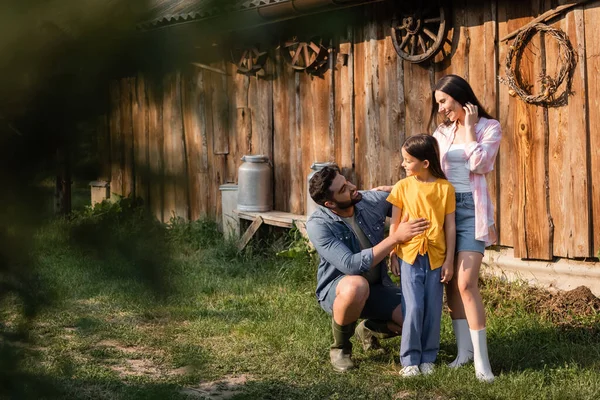 Happy man hugging daughter near smiling wife on rural farm and blurred foreground - foto de stock