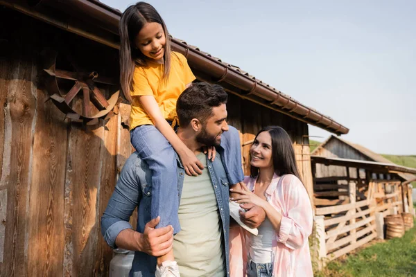 Happy man piggybacking daughter near wife and wooden barn on farm — Fotografia de Stock