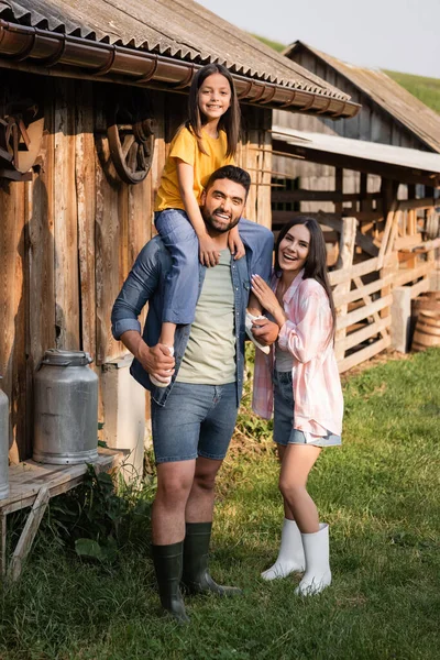 Happy man piggybacking daughter near laughing wife on farm in village - foto de stock