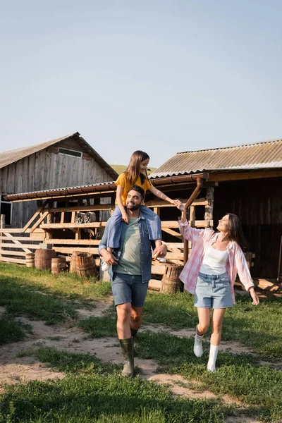 Cheerful man piggybacking daughter while walking near wife on rural farm — Stock Photo