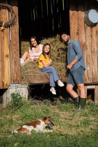 Happy couple looking at camera near haystack in barn and dog on foreground - foto de stock