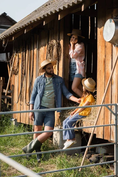 Girl in straw hat sitting on hay in barn near happy parents — Photo de stock