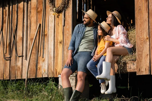 Family of farmers in straw hats sitting on hay in wooden barn and looking away — Foto stock