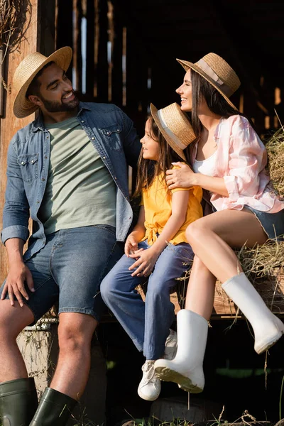 Cheerful couple in straw hats looking at each other near daughter on hay in barn — Foto stock