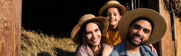 Granjeros alegres con hija en sombrero de paja sonriendo a la cámara cerca del heno en la granja, pancarta - foto de stock