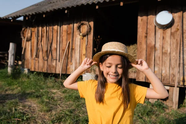 Smiling child with closed eyes touching straw hat near wooden barn on blurred background — Stockfoto