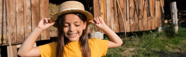 Cheerful girl with closed eyes adjusting straw hat on farm in village, banner — Stock Photo