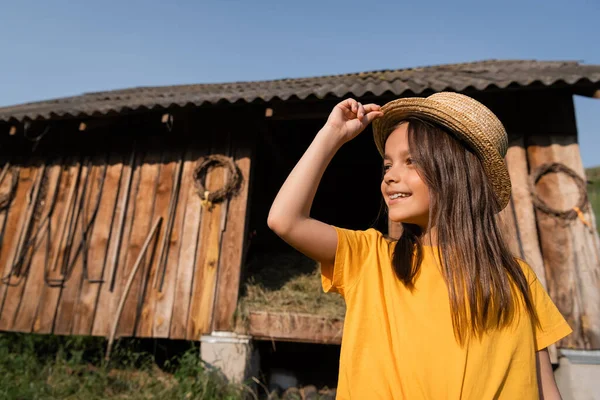 Chica feliz en sombrero de paja mirando lejos cerca de granero de madera en la granja del pueblo - foto de stock