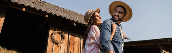 Mulher feliz em chapéu de palha abraçando marido barbudo perto de celeiro de madeira, bandeira — Stock Photo