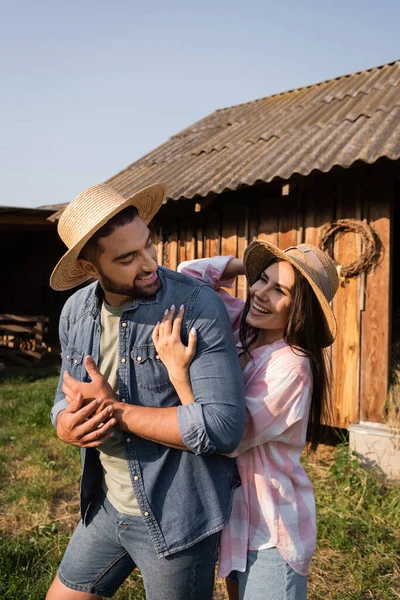 Mujer alegre en sombrero de paja abrazando marido en granja en el campo - foto de stock