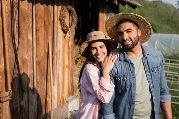 Granjeros satisfechos en sombreros de paja mirando a la cámara cerca de granero de madera en el día de verano - foto de stock