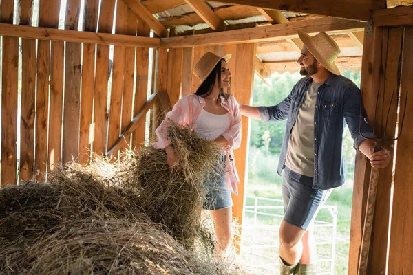 Bearded farmer in straw hat talking to smiling wife stacking hey in barn — Photo de stock