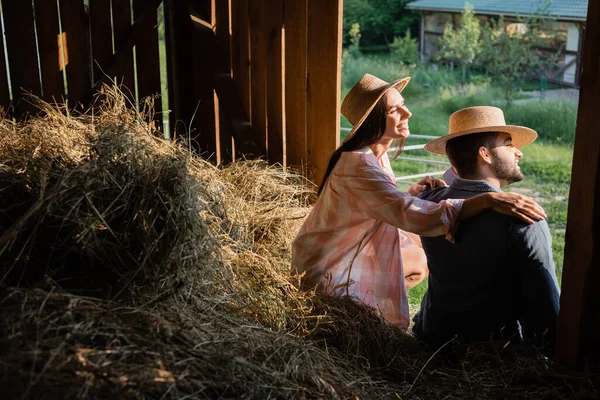 Happy woman in straw hat hugging shoulders of husband while sitting on haystack in barn — Foto stock