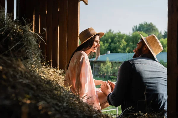 Счастливые фермеры в соломенных шляпах держатся за руки и смотрят друг на друга в сарае рядом с эй — стоковое фото