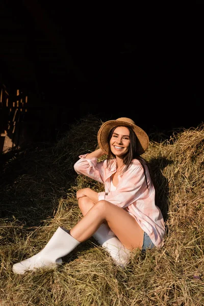 Mujer granja feliz en sombrero de paja sentado en el pajar y mirando a la cámara - foto de stock