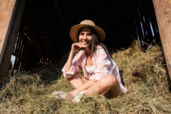 Довольная женщина в соломенной шляпе сидит на стоге сена со скрещенными ногами и смотрит в камеру — стоковое фото
