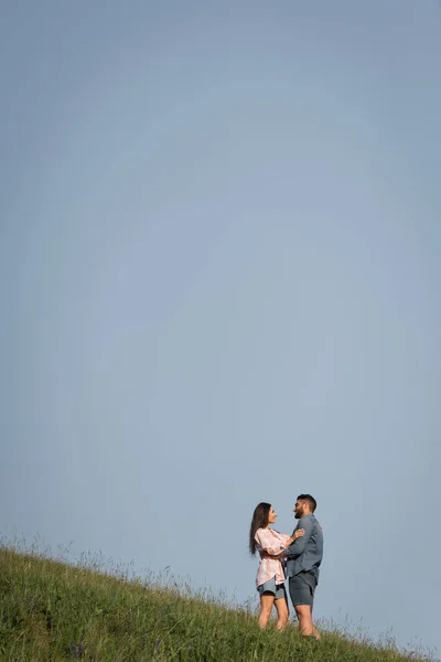Feliz pareja de pie cara a cara y abrazándose en la pendiente bajo el cielo azul - foto de stock