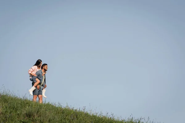 Uomo barbuto piggybacking donna in stivali di gomma sul pendio verde — Foto stock