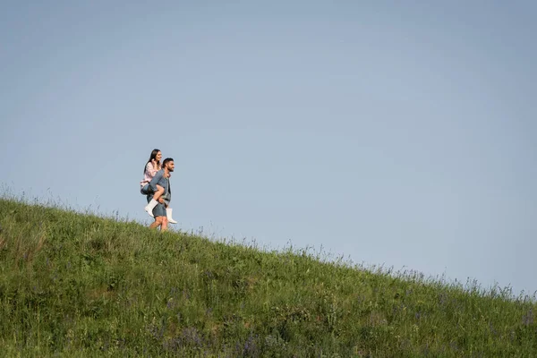 Vista da lontano su uomo piggybacking fidanzata sul pendio erboso — Foto stock