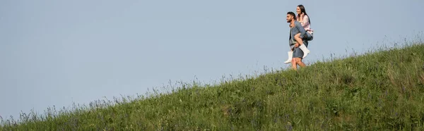 View from afar on man piggybacking woman in hilly meadow under blue sky, banner — Fotografia de Stock