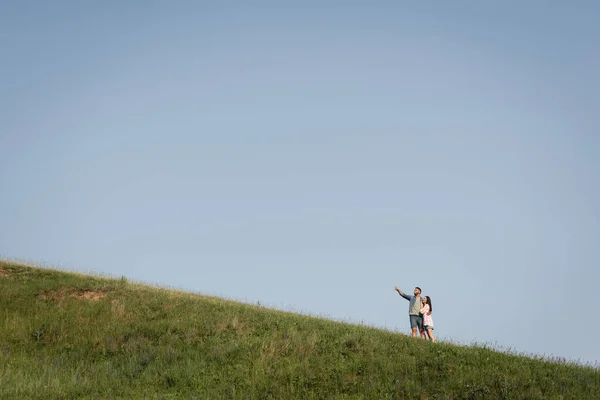 View from afar on man embracing woman and pointing with finger in green meadow — Stockfoto