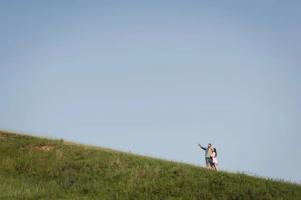 Blick aus der Ferne auf Mann, der Frau umarmt und mit Finger auf grüne Wiese zeigt — Stockfoto